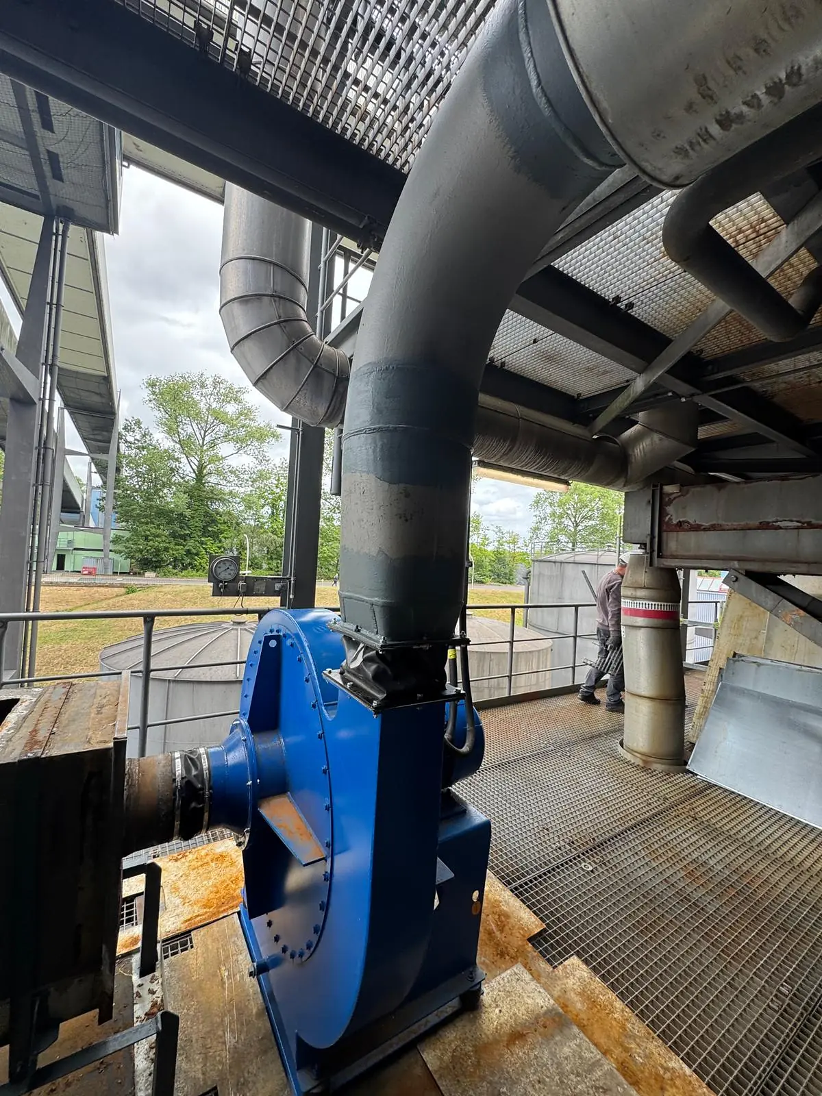New blue centrifugal fan installed on a steel base on an outdoor grating platform, its outlet connected through an expansion joint to the existing painted exhaust stack