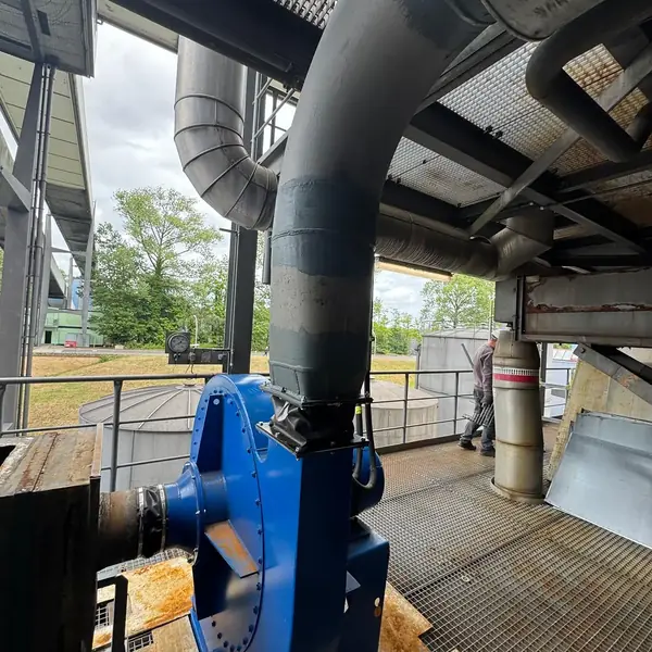New blue centrifugal fan installed on a steel base on an outdoor grating platform, its outlet connected through an expansion joint to the existing painted exhaust stack