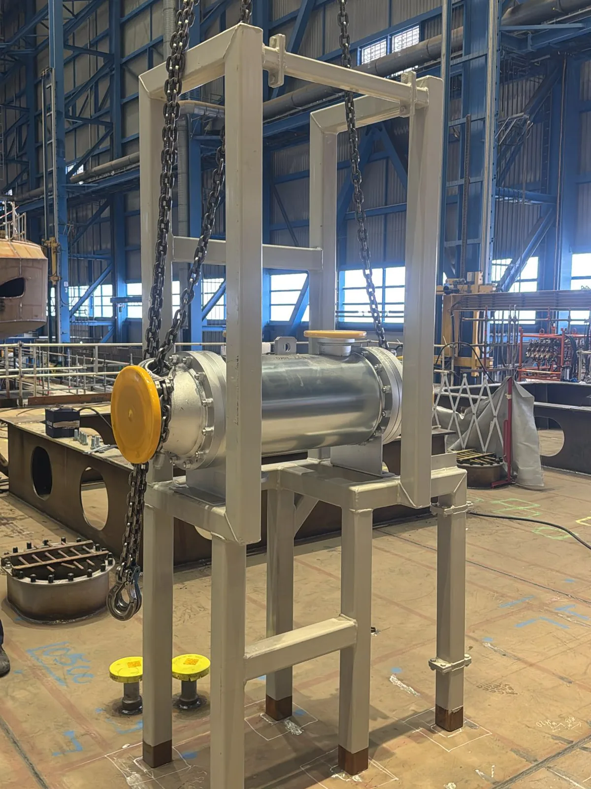 Cylindrical steel unit in a fabricated frame, suspended on chain hoists over its marked position on the deck of a vessel under construction