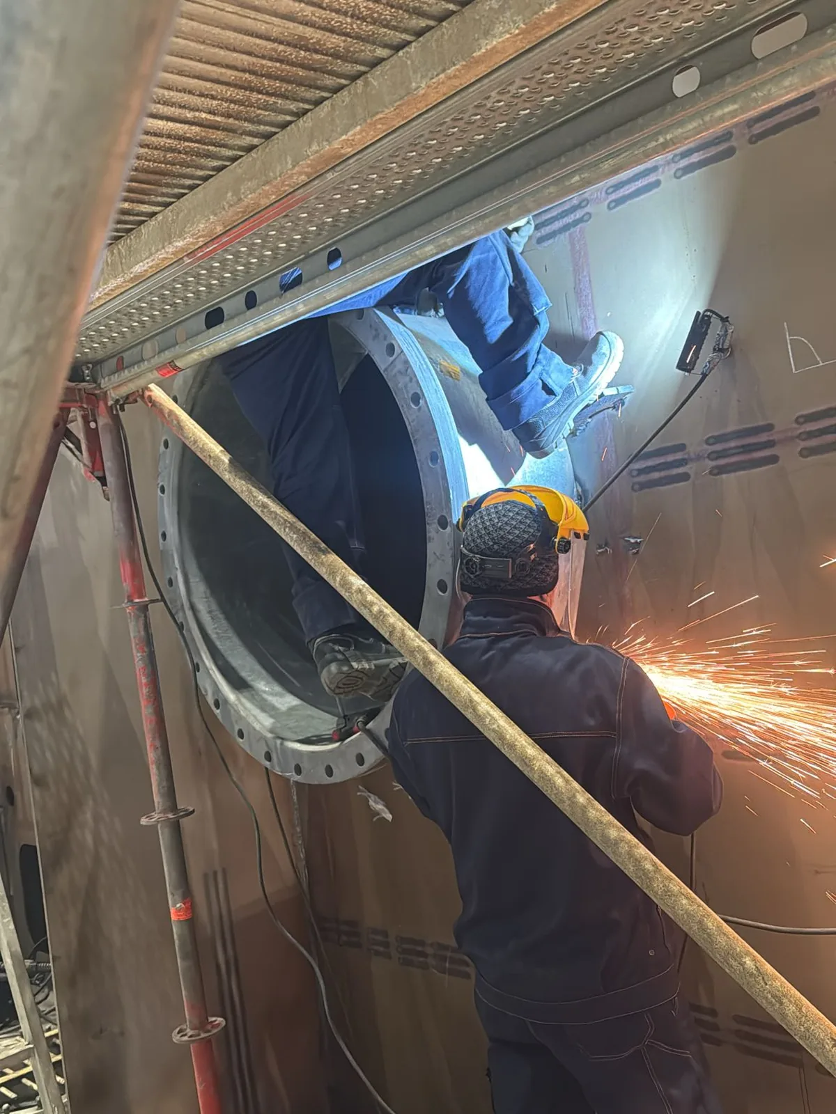 Two fitters at a bulkhead penetration, one checking alignment inside the opening while the other dresses the edge with a grinder