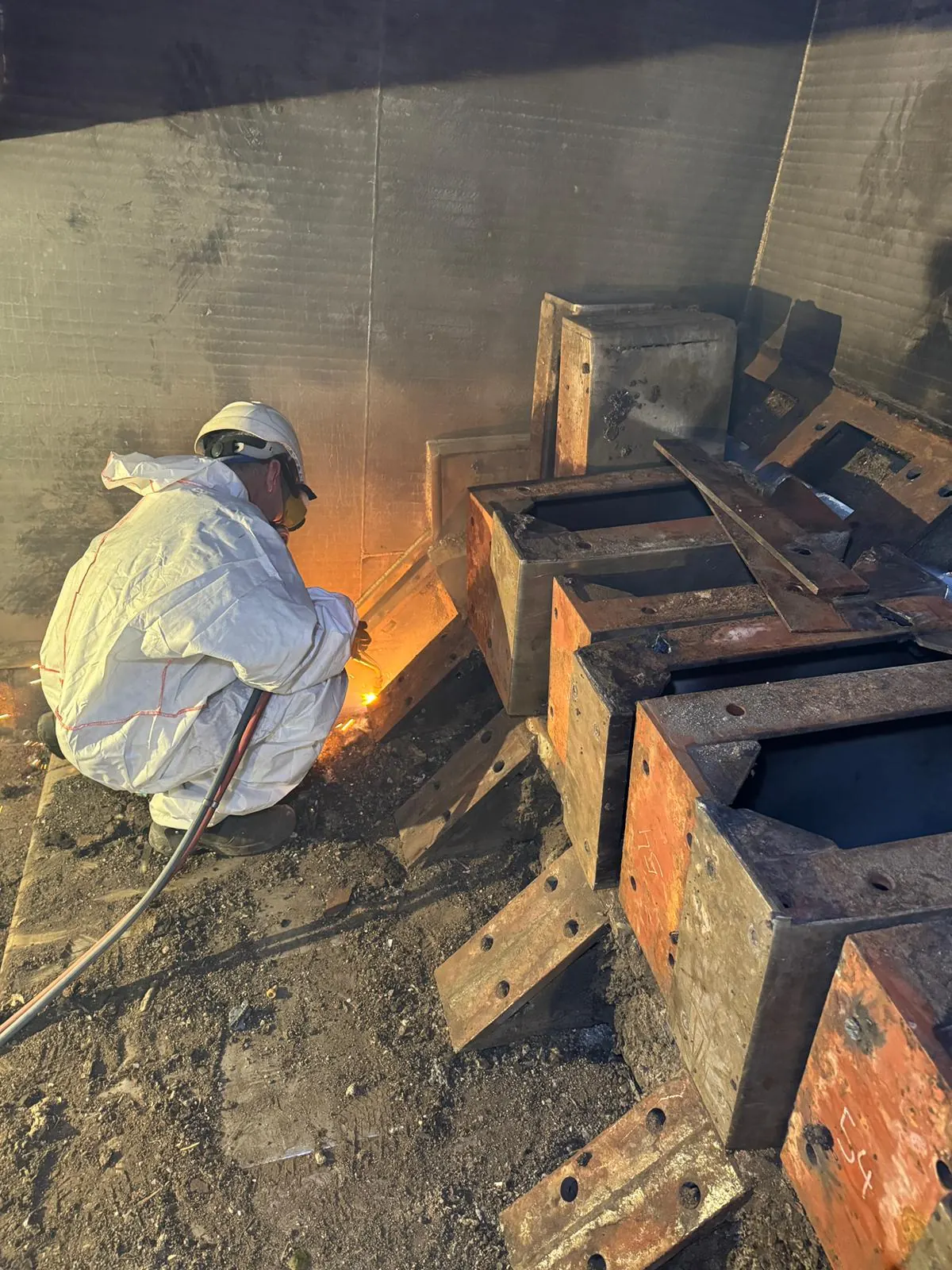 Worker in disposable coveralls and respirator cutting seized steel free with an oxy-fuel torch at the grate, with removed grate blocks in the foreground