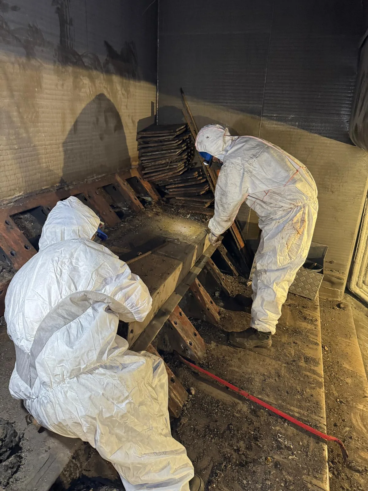 Two workers in disposable coveralls levering an old grate bar out of its row, with removed bars stacked behind them