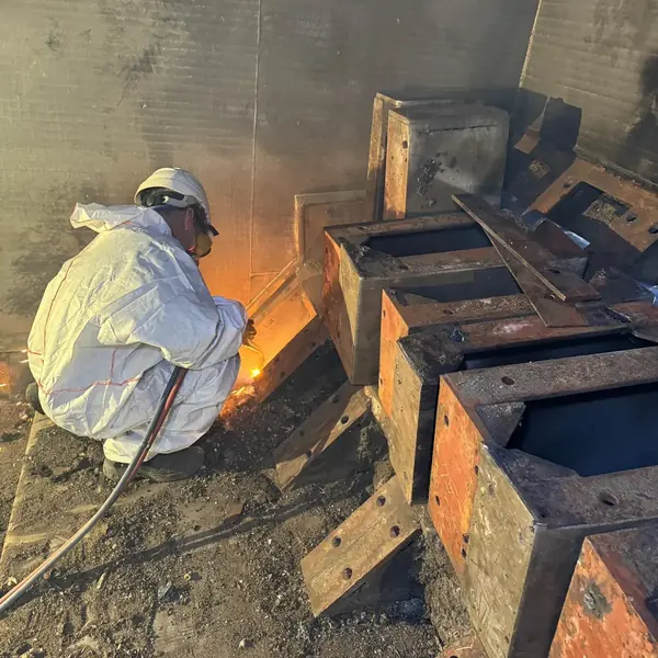 Worker in disposable coveralls and respirator cutting seized steel free with an oxy-fuel torch at the grate, with removed grate blocks in the foreground