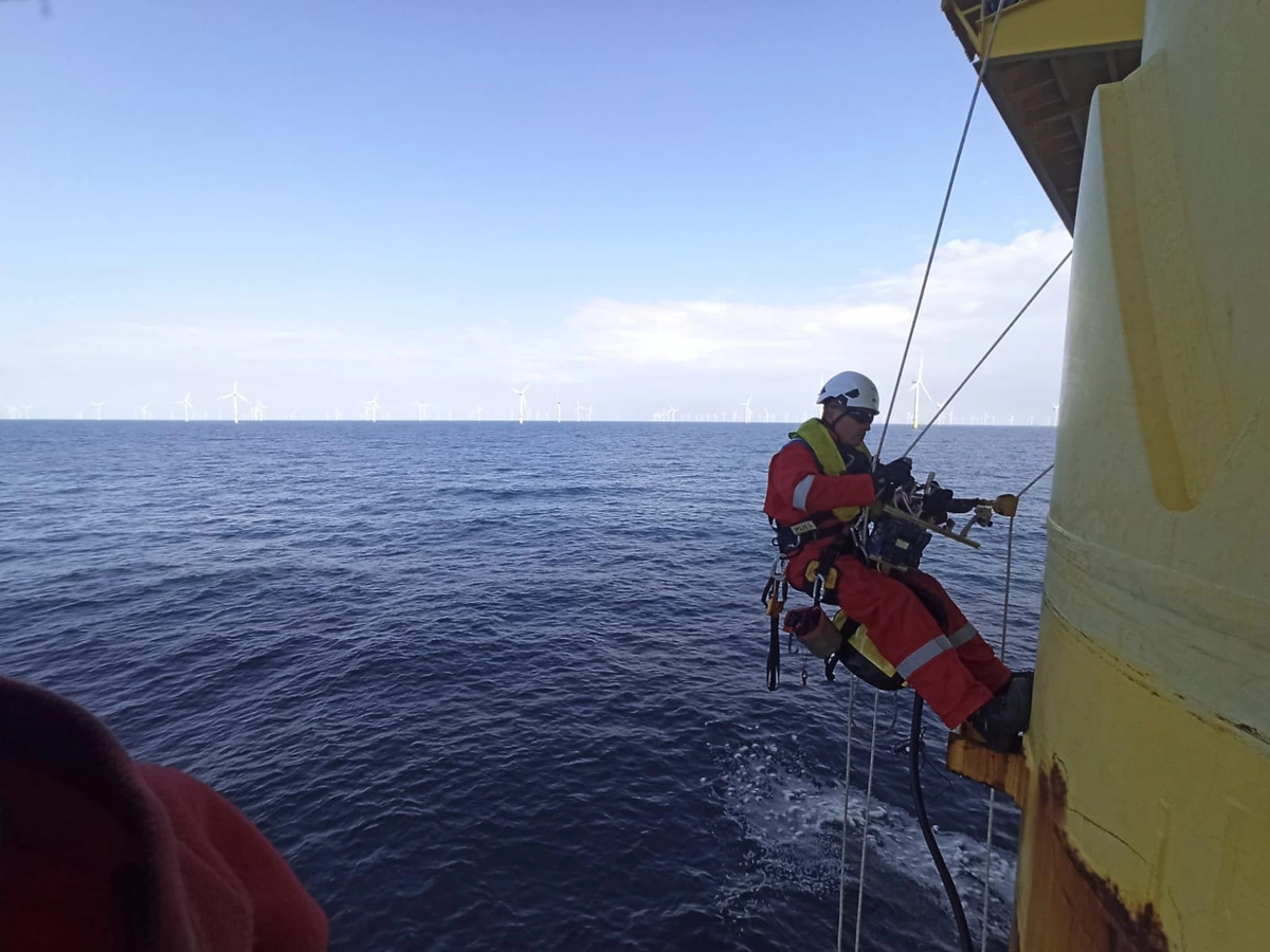 Rope access technician suspended in a harness on the outside of an offshore platform, with wind turbines on the horizon