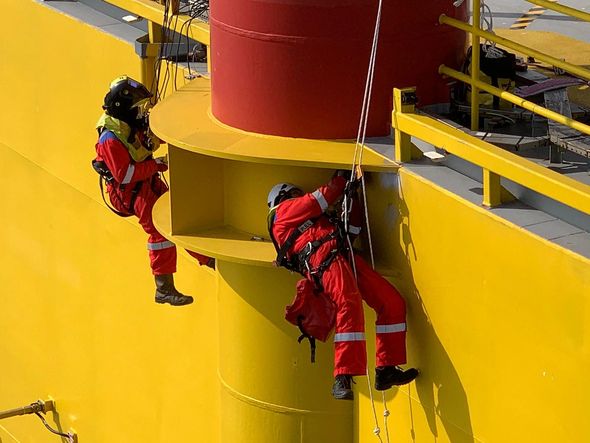 Two rope access technicians suspended on twin ropes at a yellow structure on an offshore platform, one wearing a welding helmet, with cables run down from the deck above