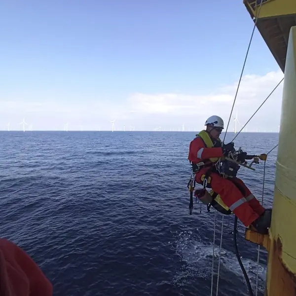 Rope access technician suspended in a harness on the outside of an offshore platform, with wind turbines on the horizon