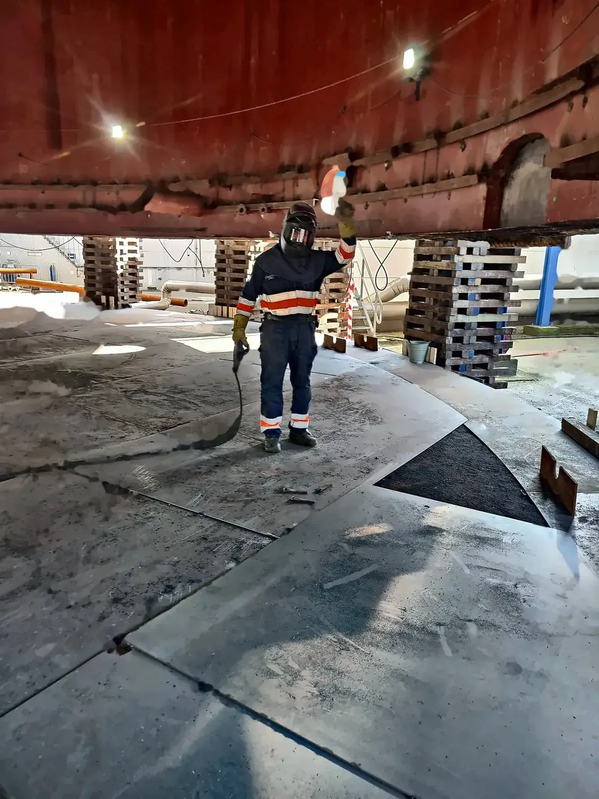 Welder working under a tank shell held up and secured on timber packing, with bottom plates laid out on the shop floor