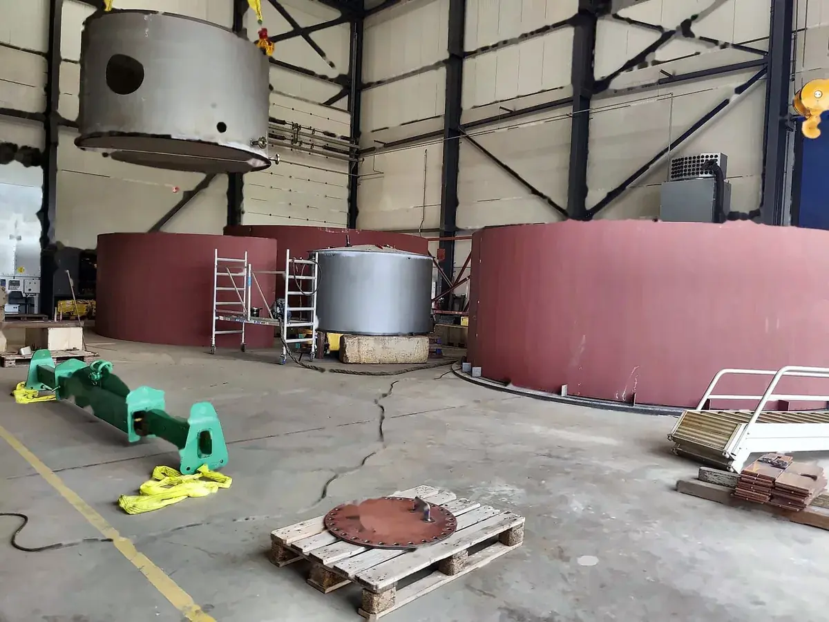 Fabrication hall with tank shells, a vessel on packing, and a spreader beam with slings on the floor