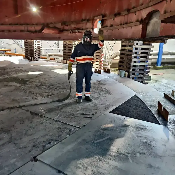 Welder working under a tank shell held up and secured on timber packing, with bottom plates laid out on the shop floor